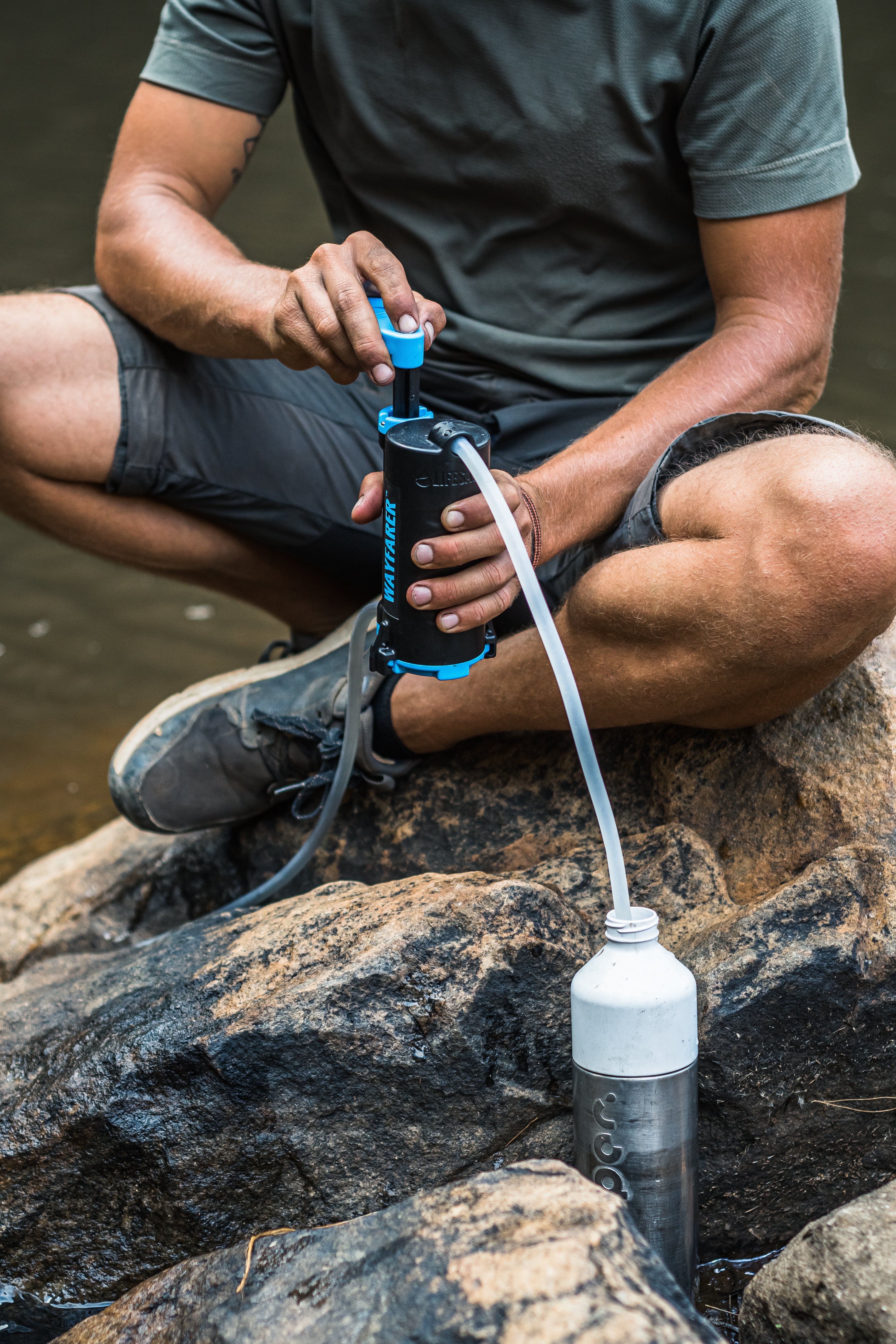 A man crouching on rocks by a body of water, using a LifeSaver Wayfarer to fill a clear water bottle with a clear hose.