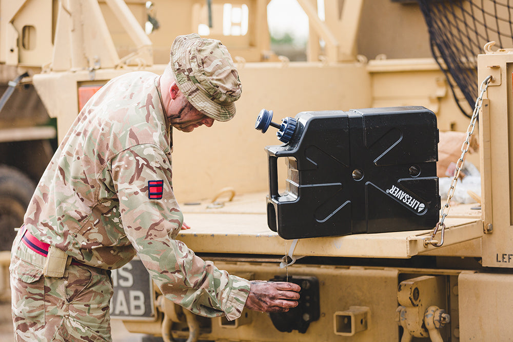 A soldier in camouflage uniform works beside a black LifeSaver Jerrycan mounted on a tan military vehicle.