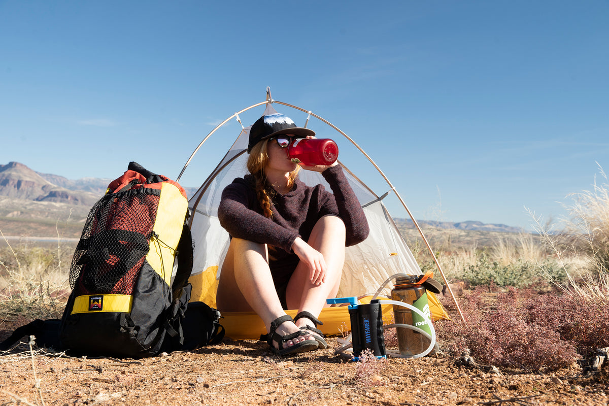 A woman sits on the ground in front of a small dome tent, drinking from a red water bottle. A large yellow, black, and red hiking backpack stands to her left, with aLifeSaver Wayfarer in front of her.