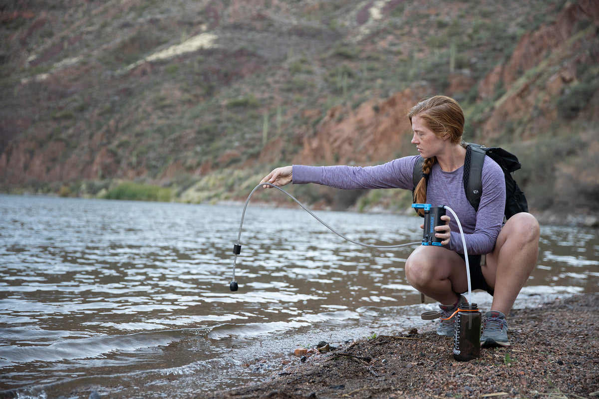 A woman crouches by a lake or riverbank, using a LifeSaver Wayfarer connected to a hose. She is wearing a purple long-sleeve shirt, gray shorts, and a black backpack, with a rocky, desert-look hillside in the background.